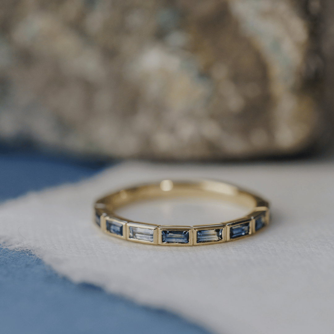 A 14k gold ring featuring blue baguette-cut sapphire gemstones displayed on a white surface with a soft-focus background.