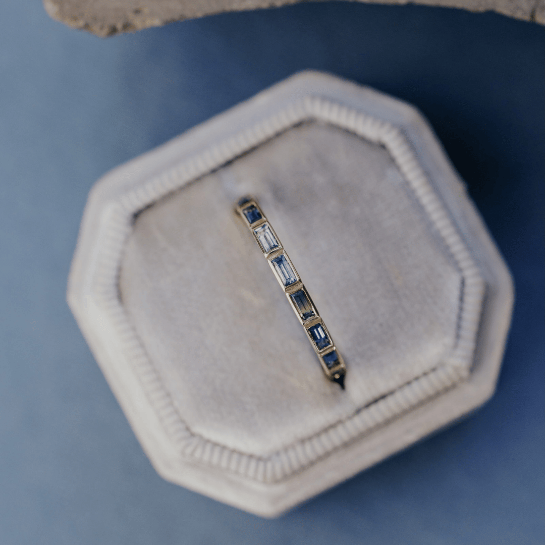 A 14k gold ring featuring blue baguette-cut sapphire gemstones from a top view, displayed in a ring box with a soft-focus background.
