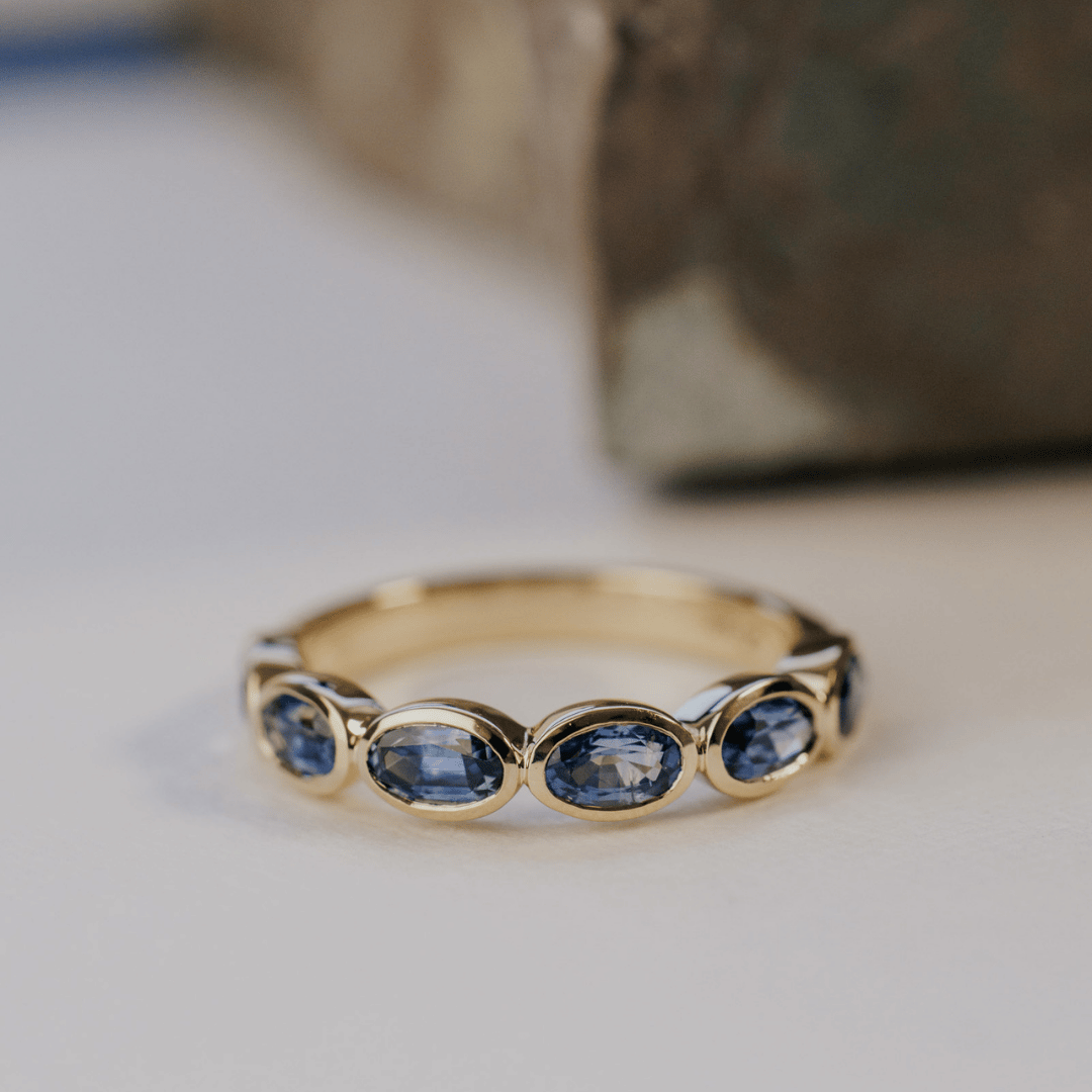 A 14k gold ring featuring blue oval sapphire gemstones displayed on a white surface with a soft-focus background.