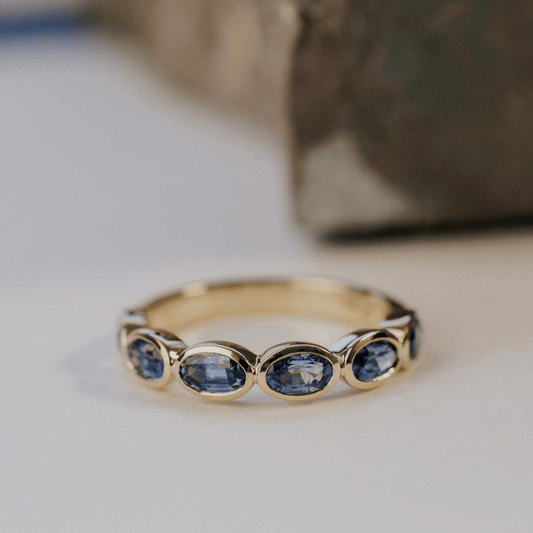 A 14k gold ring featuring blue oval sapphire gemstones displayed on a white surface with a soft-focus background.
