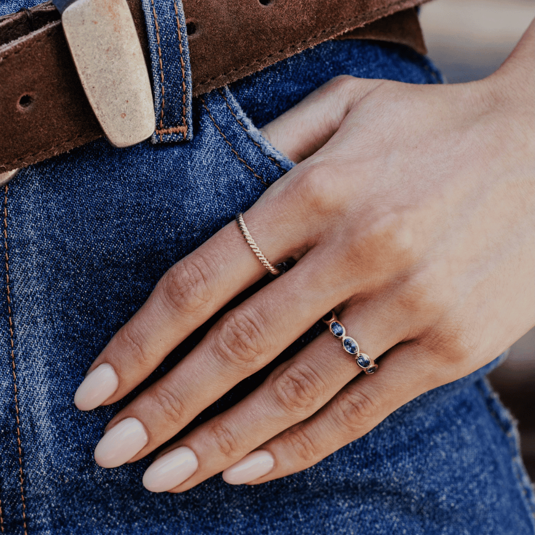 A 14k gold ring featuring blue oval sapphire gemstones shown on the ring finger of a hand model. 