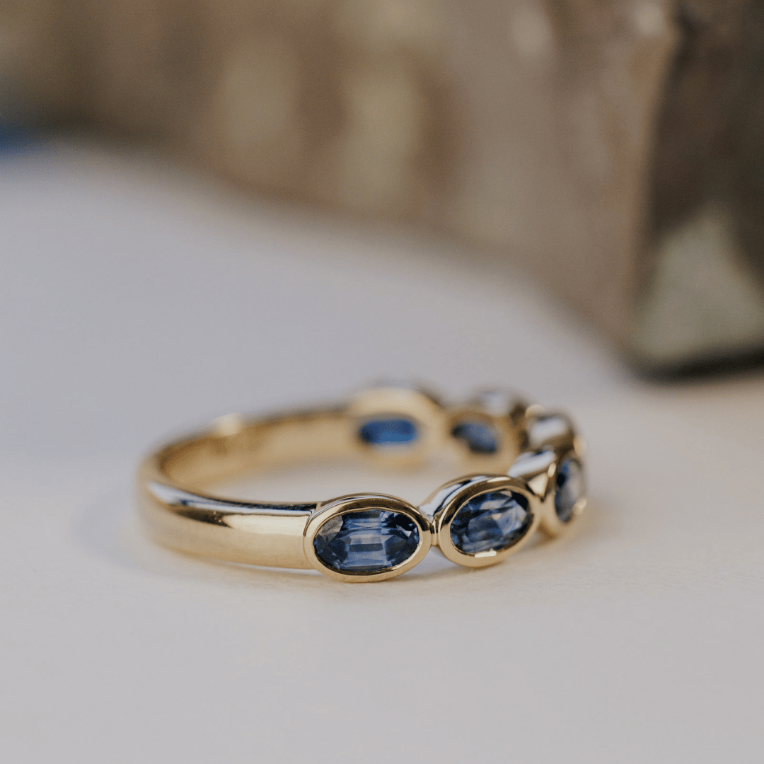 A 14k gold ring featuring blue oval sapphire gemstones from an angled view, displayed on a white surface with a soft-focus background.