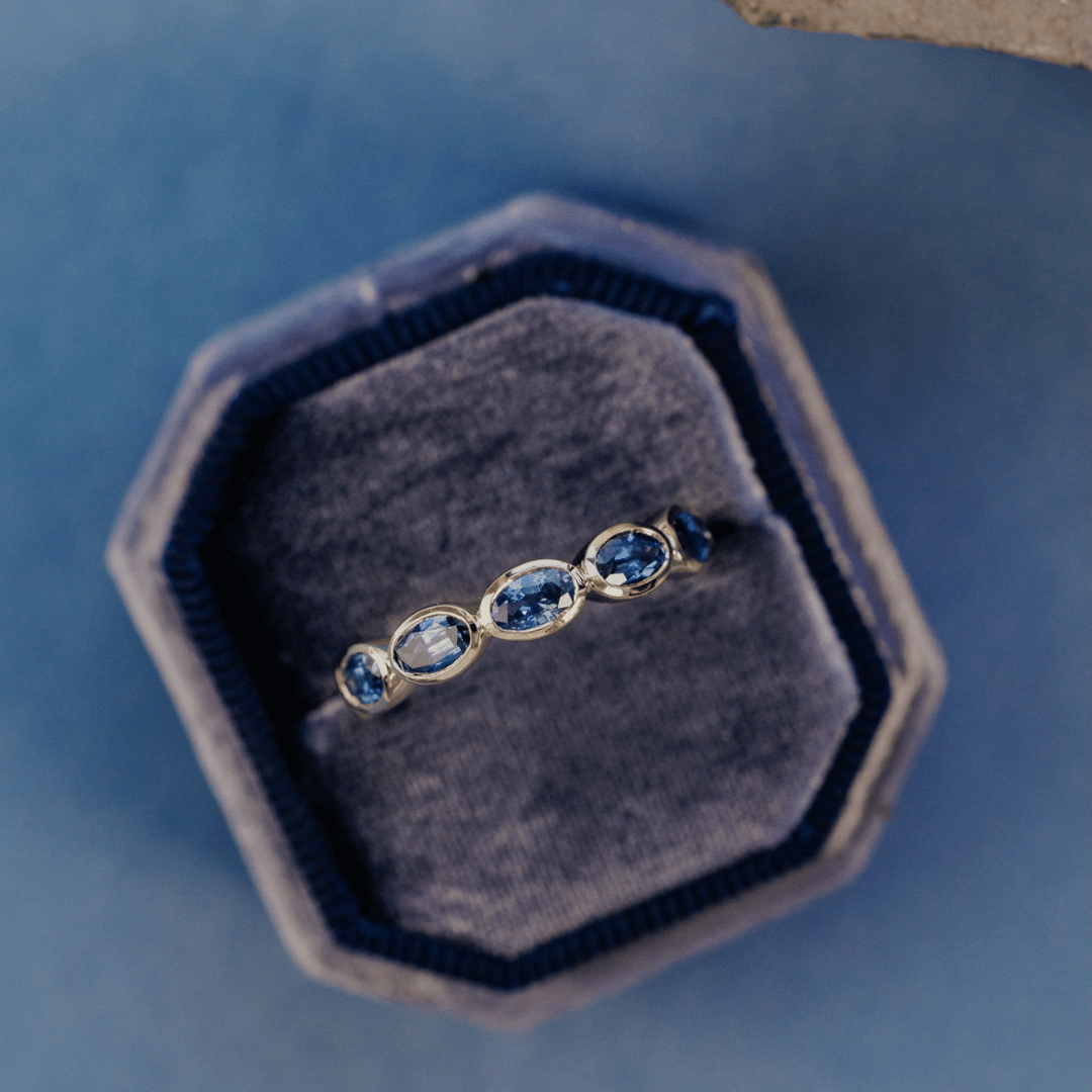 A 14k gold ring featuring blue oval sapphire gemstones from a top view, displayed in a ring box with a soft-focus background.