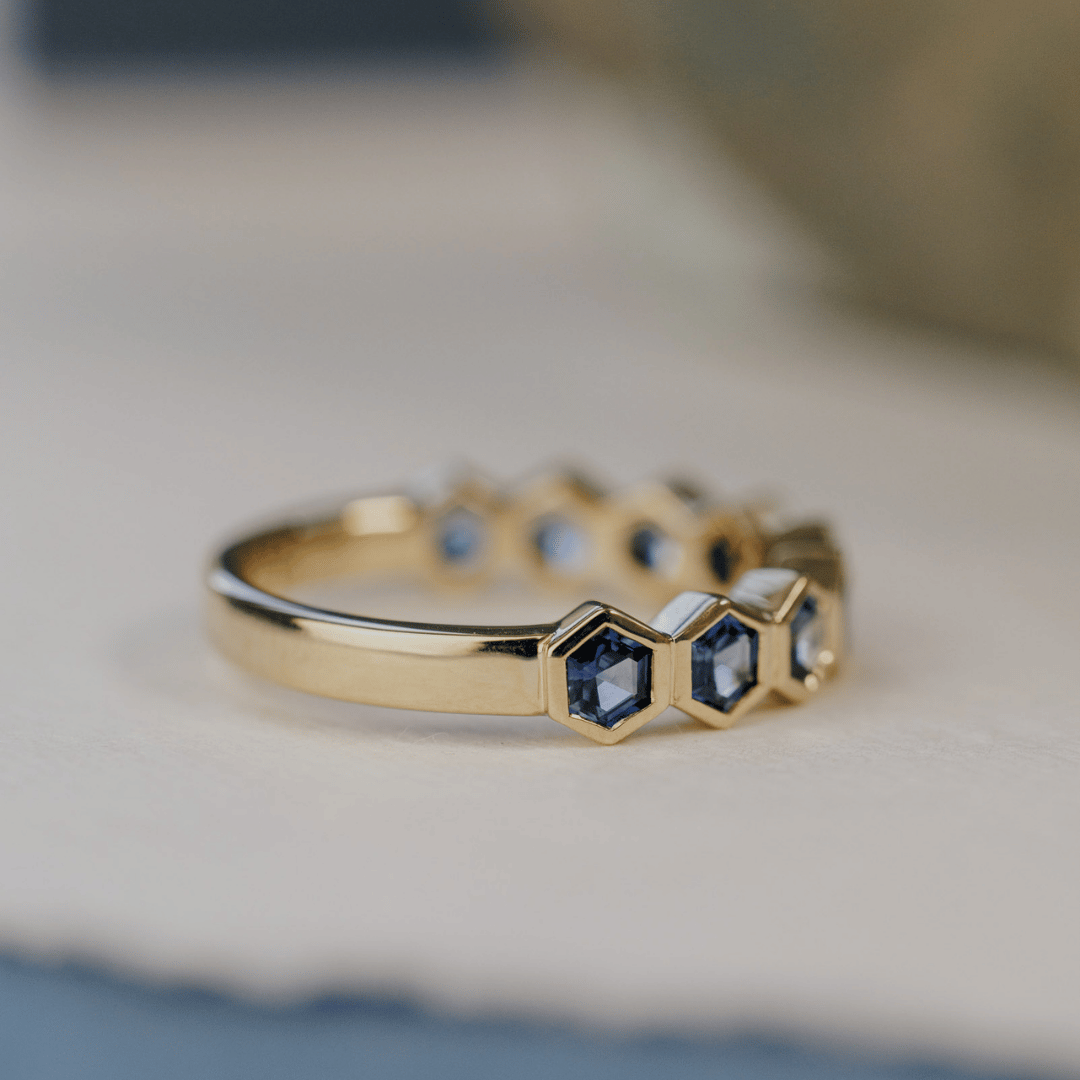 A 14k gold ring featuring blue hexagonal cut sapphire gemstones from an angled view, displayed on a white surface with a soft-focus background.