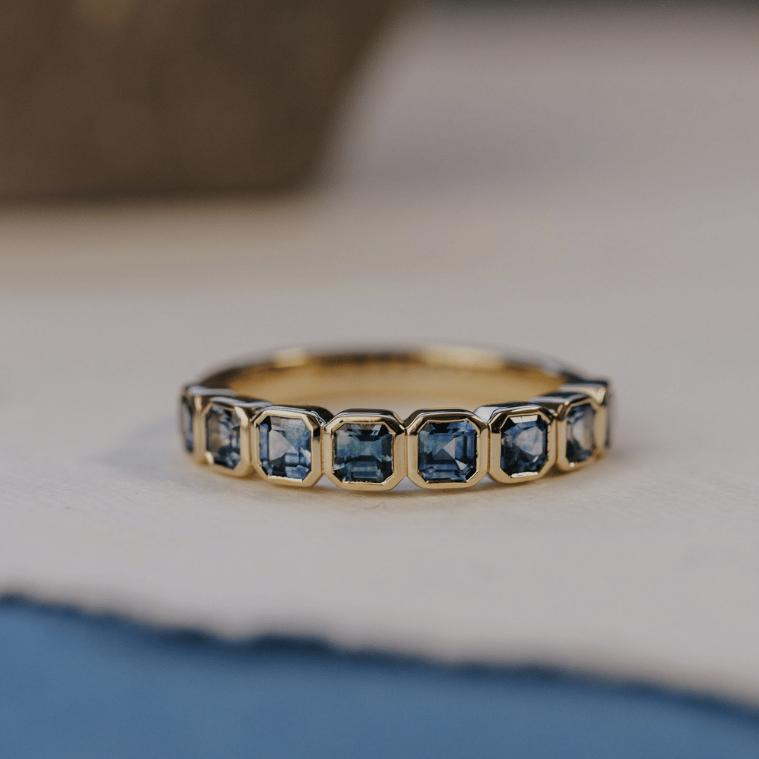 A 14k gold ring featuring a asscher cut, blue sapphire gemstones, displayed on a white surface with a soft-focus background.