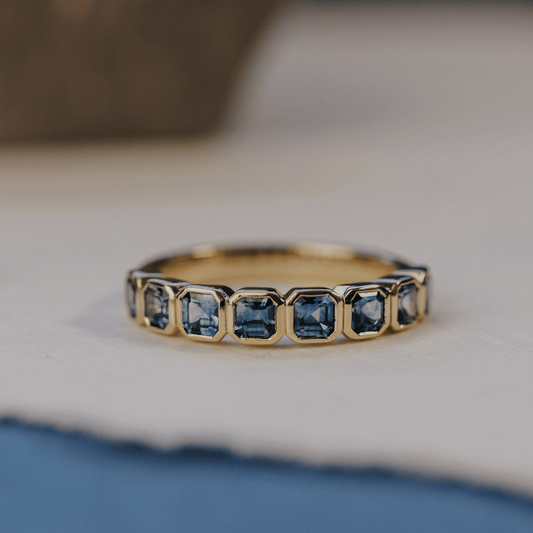 A 14k gold ring featuring a asscher cut, blue sapphire gemstones, displayed on a white surface with a soft-focus background.
