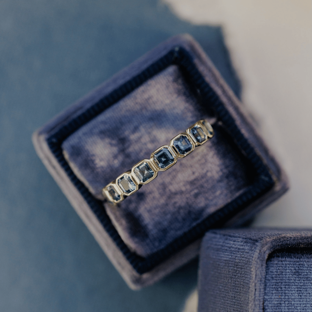 A 14k gold ring featuring a asscher cut, blue sapphire gemstones, displayed in a ring boxon a blue surface with a soft-focus background.