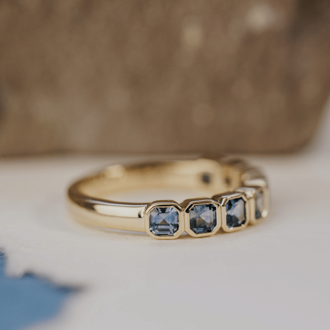 A 14k gold ring featuring asscher cut, blue sapphire gemstones, set to the side and displayed on a white surface with a soft-focus background.