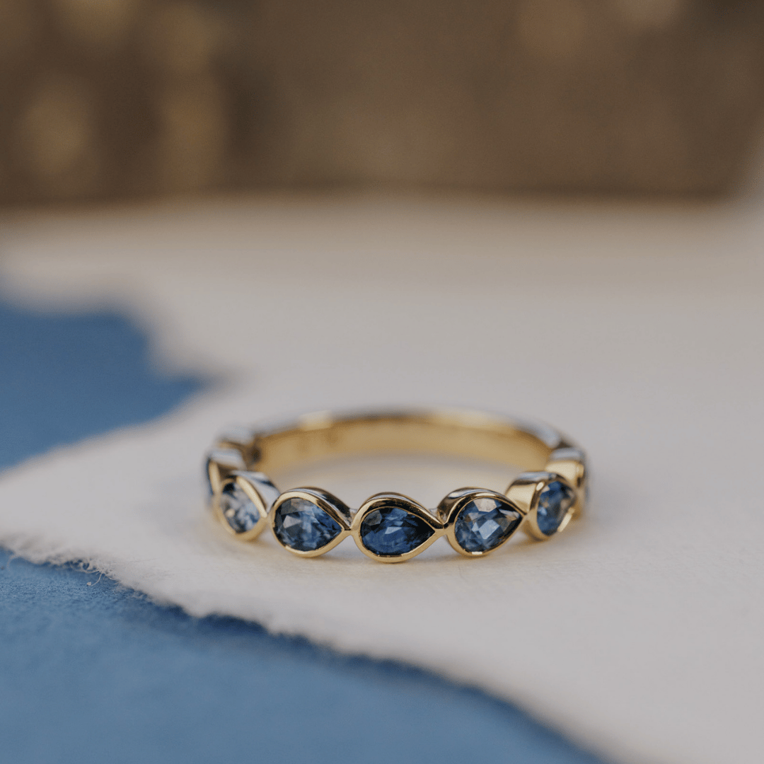 A 14k gold ring featuring blue pear sapphire gemstones displayed on a white surface with a soft-focus background.