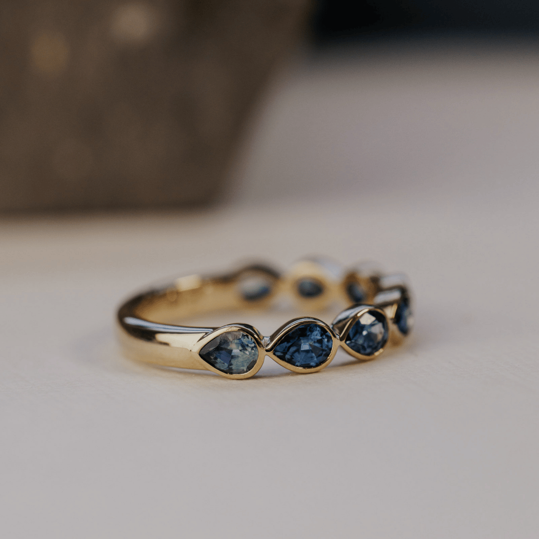 A 14k gold ring featuring blue pear sapphire gemstones displayed at an angle on a white surface with a soft-focus background.