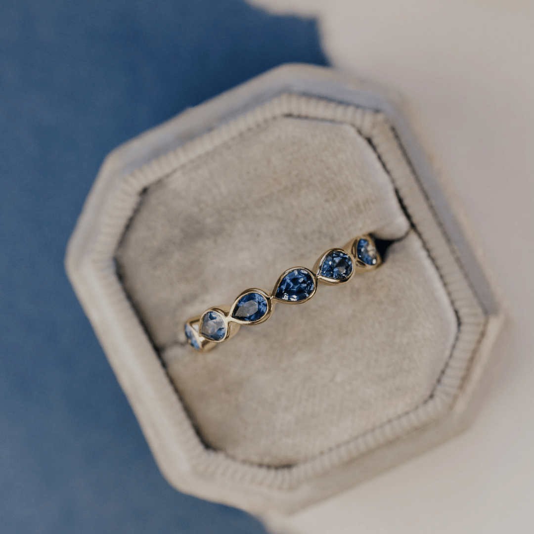 A 14k gold ring featuring blue pear sapphire gemstones displayed in a velvet ring box on a white and blue surface with a soft-focus background.