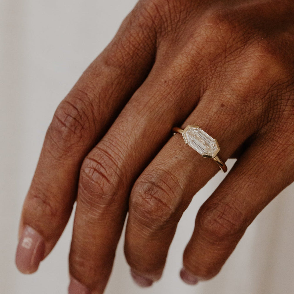 Hand wearing a gold ring with a lab grown diamond stone on a neutral background.
