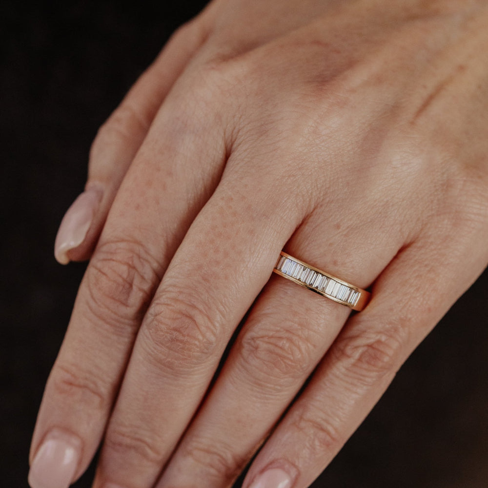 Close-up of a hand wearing a rose gold wedding band with baguette diamonds on a dark background.