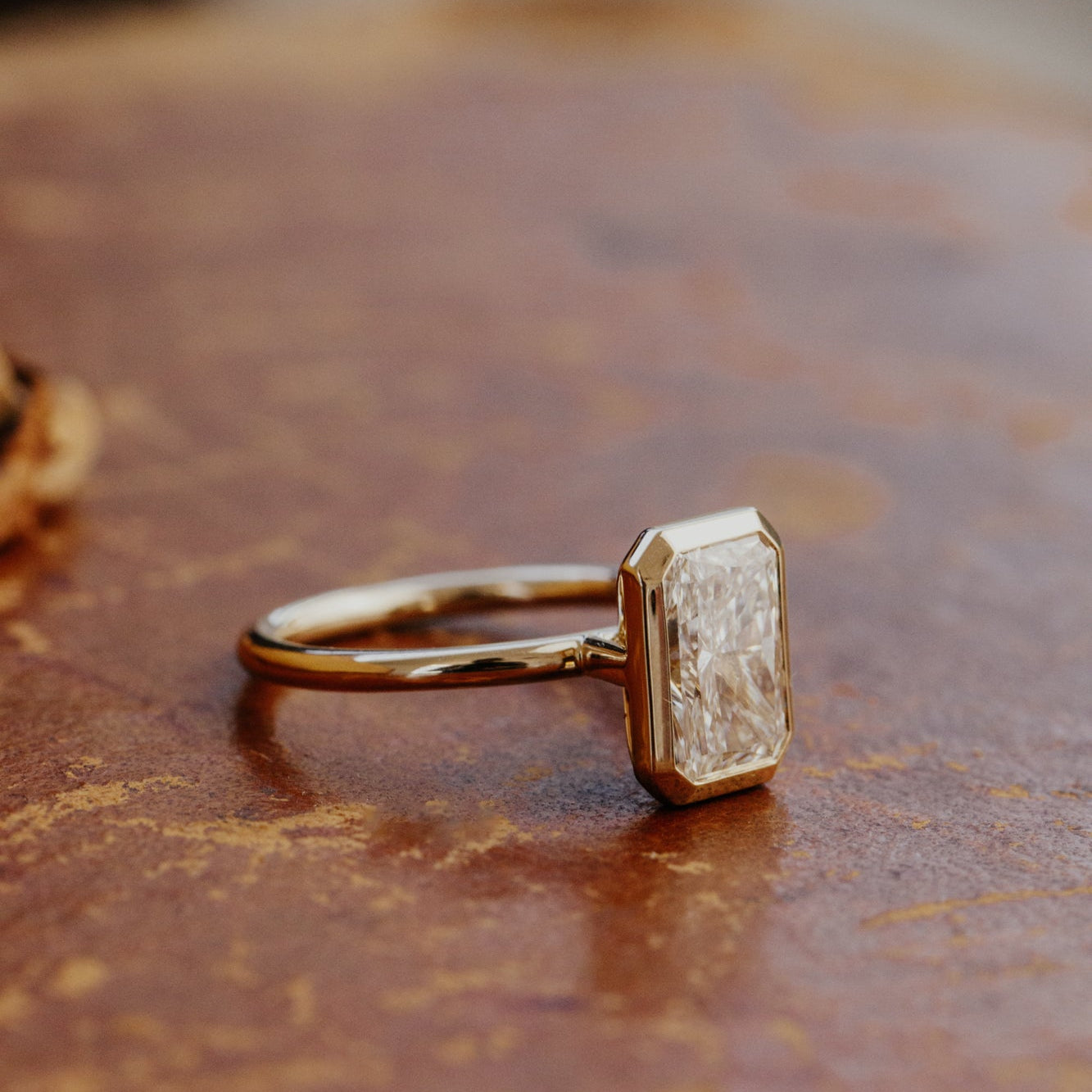 Gold Bailey bezel set  engagement ring with a radiant cut diamond on a brown surface.