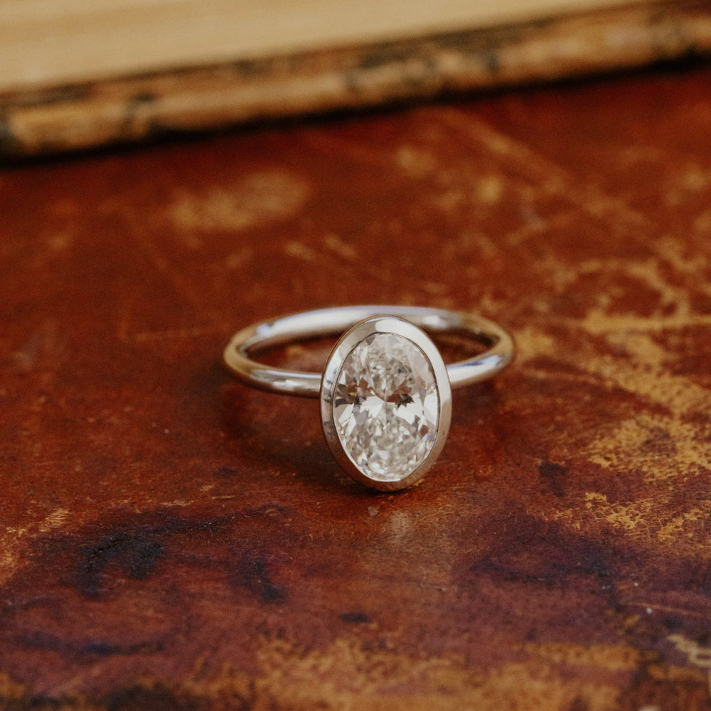 The Bailey bezel set white gold ring with a oval lab grown diamond on a textured brown surface.