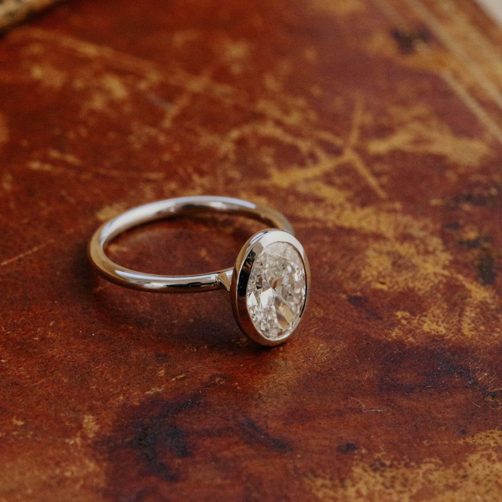 The Bailey bezel set white gold ring with a oval lab grown diamond on a textured brown surface.