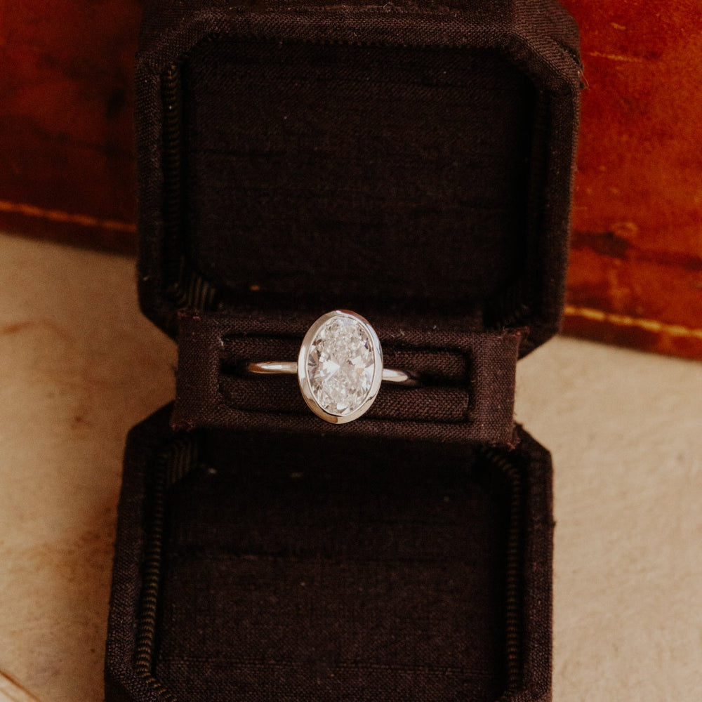 The Bailey bezel set white gold ring with a oval lab grown diamond in a black velvet box on a wooden surface.