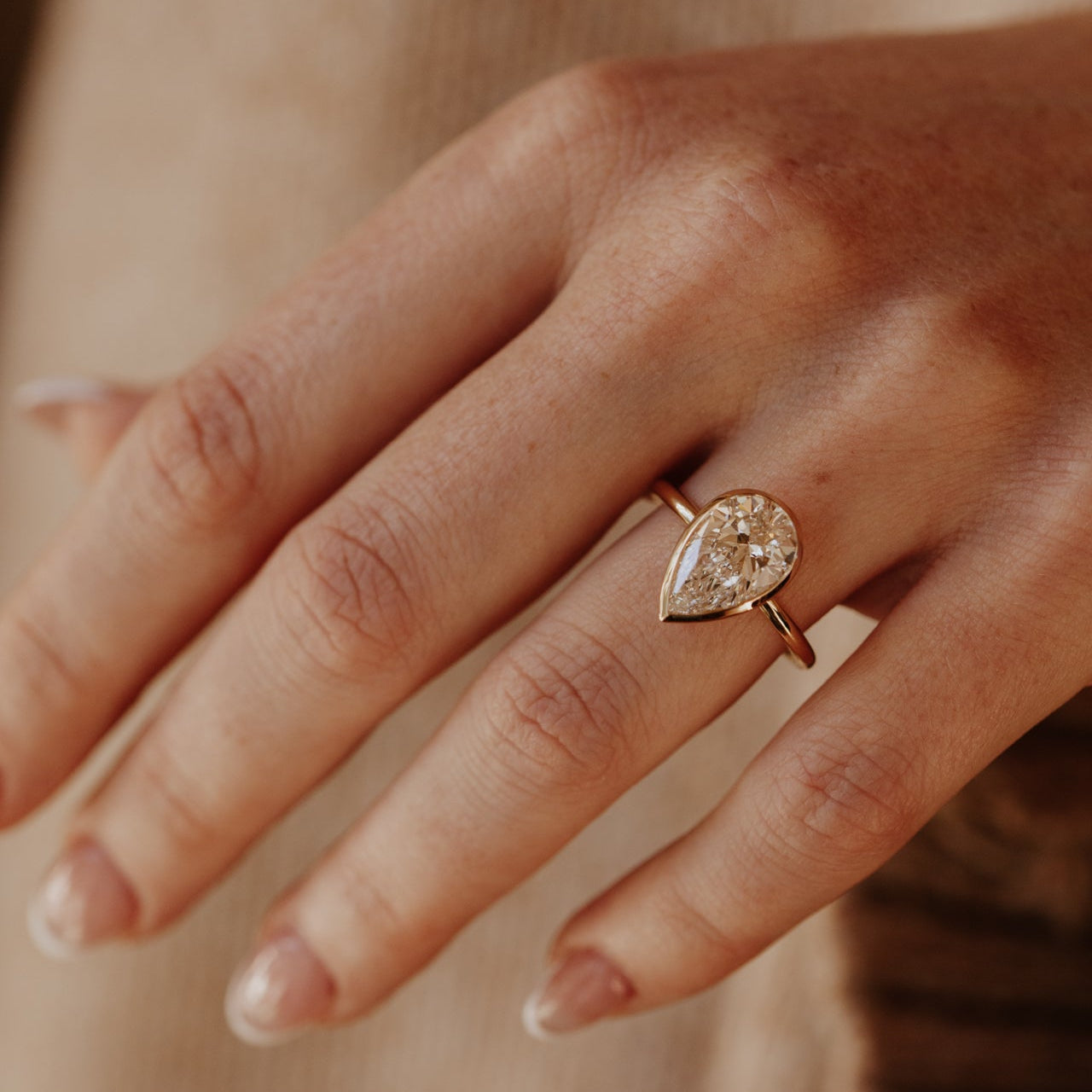 Close-up of a hand wearing the Bailey pear-shaped diamond ring set in a bezel on a blurred background.
