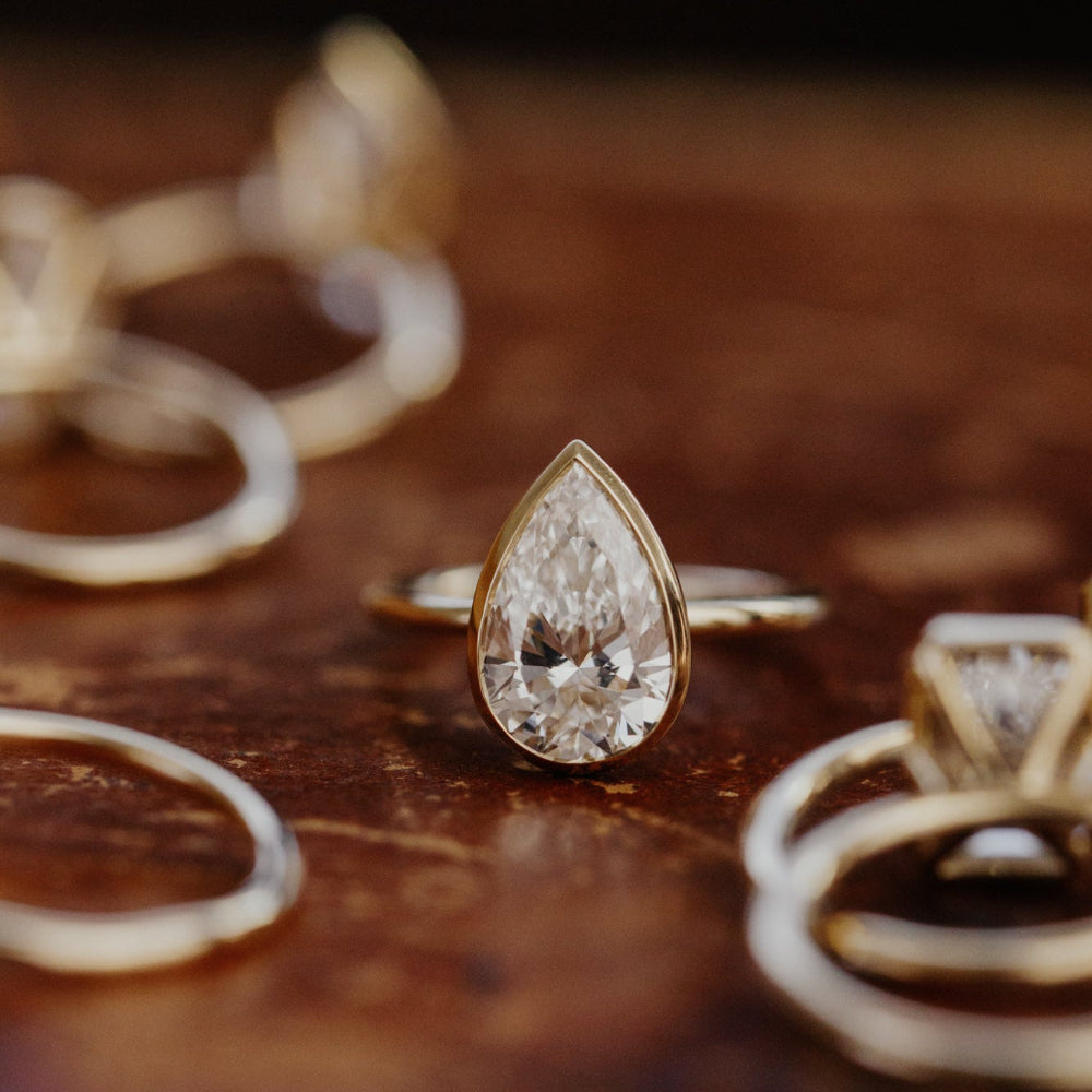 The Bailey bezel set ring with a pear-shaped diamond on a wooden surface.