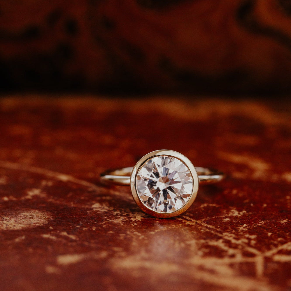 The Bailey round bezel set diamond ring on a wooden surface.