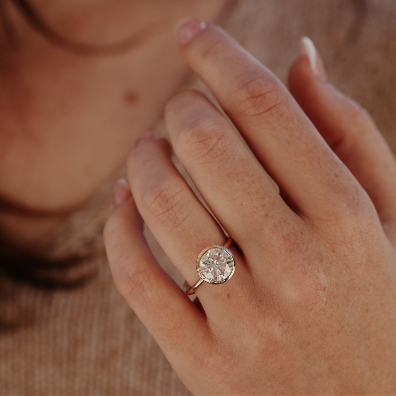 Close-up of a hand wearing the Bailey bezel ring with a round diamond on a blurred background.