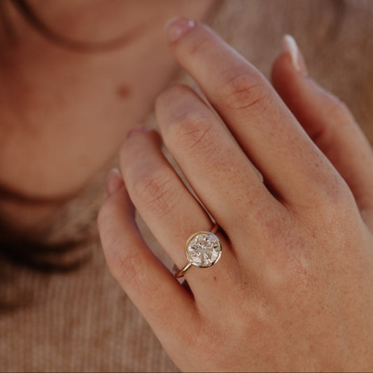 Close-up of a hand wearing the Bailey bezel ring with a round diamond on a blurred background.