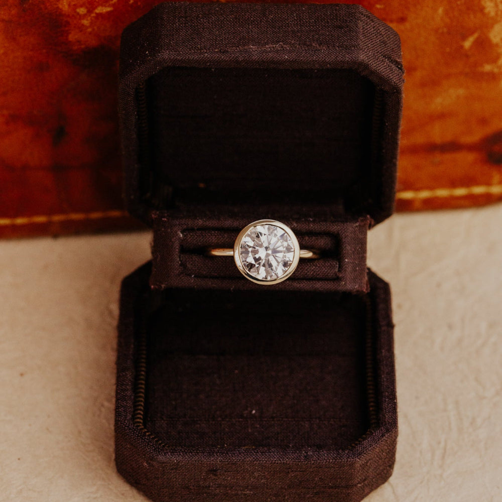 Bailey round diamond bezel set ring in a black velvet box on a textured surface with a wooden background.