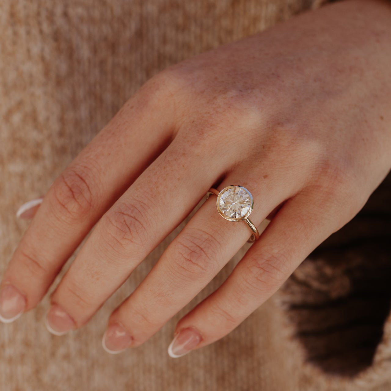 Close-up of a hand wearing the bailey bezel set ring with a round diamond on a textured background.