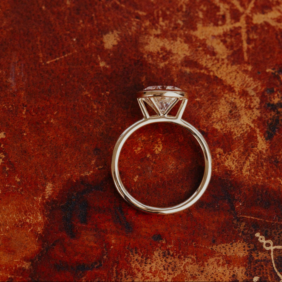 The Bailey bezel set ring with a round diamond on a textured red-brown surface.