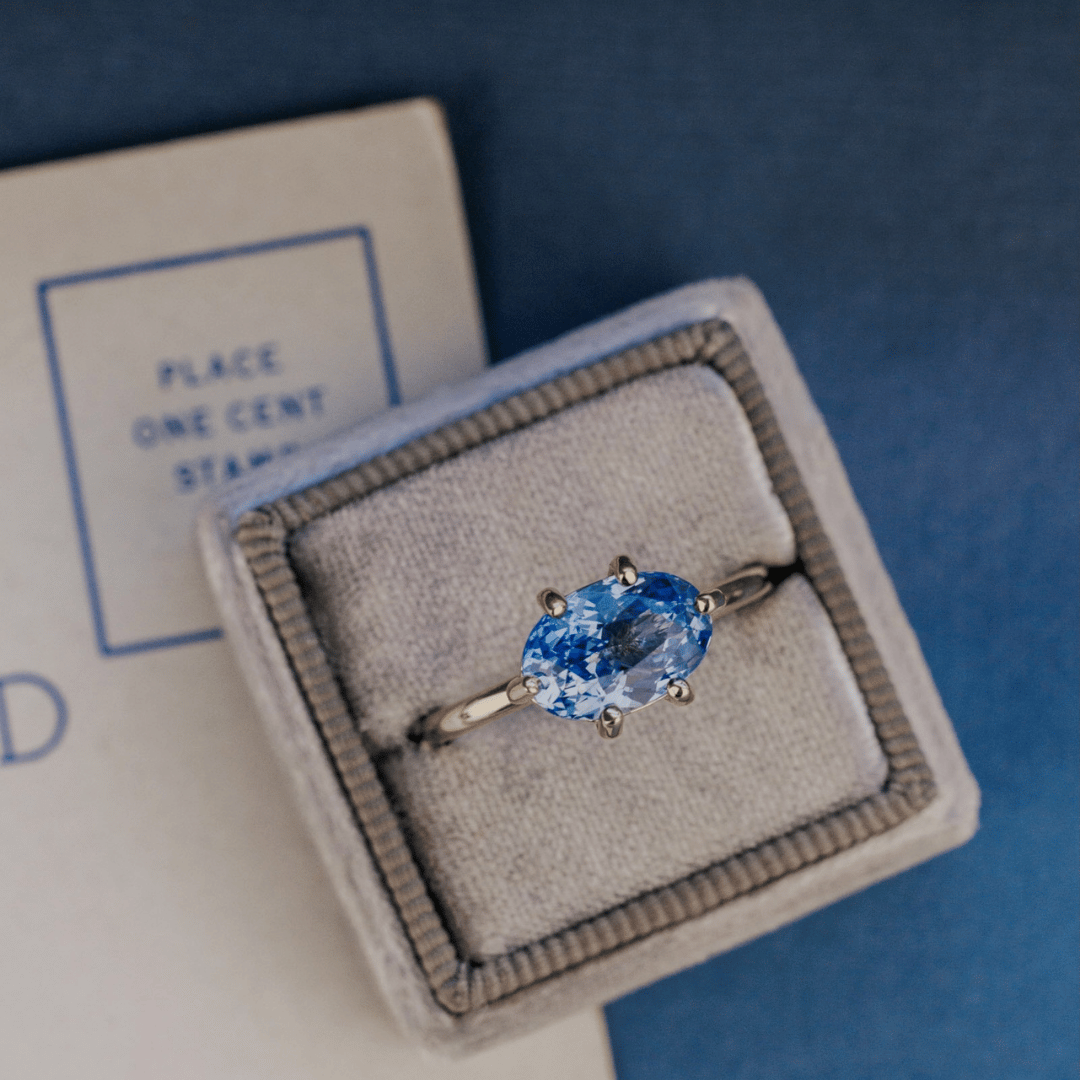 A 14k gold ring featuring a blue sapphire gemstone viewed from a ring box, displayed on a textured white and blue surface with a soft-focus background.
