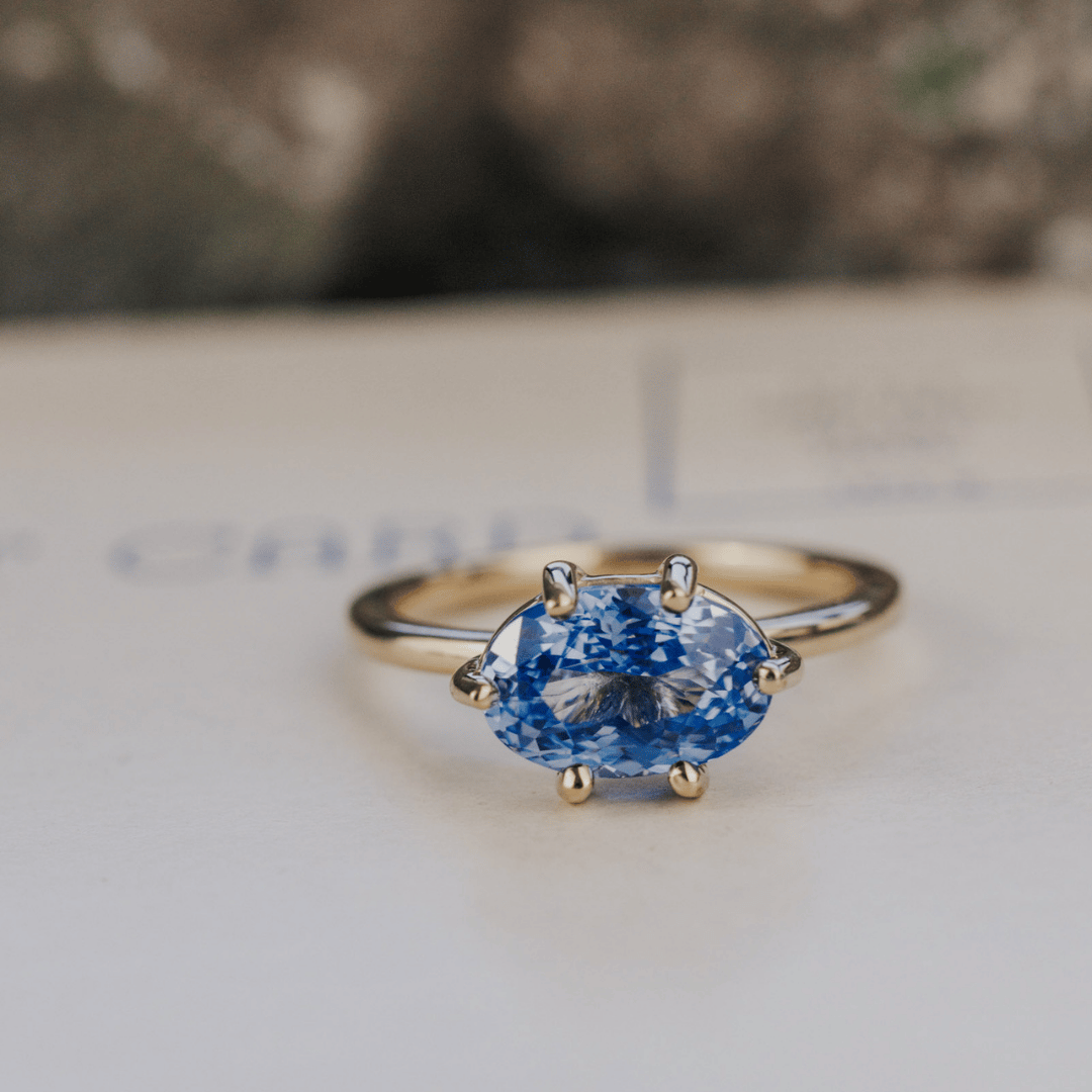 A 14k gold ring featuring a blue sapphire gemstone viewed from straight on, displayed on a textured white surface with a soft-focus background.