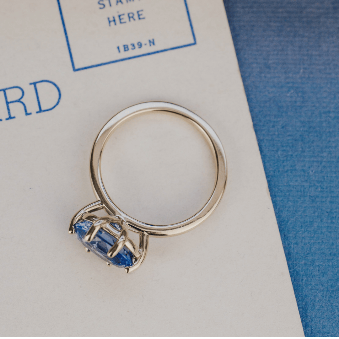A 14k gold ring featuring a blue sapphire gemstone viewed from the top, displayed on a textured white surface with a soft-focus background.