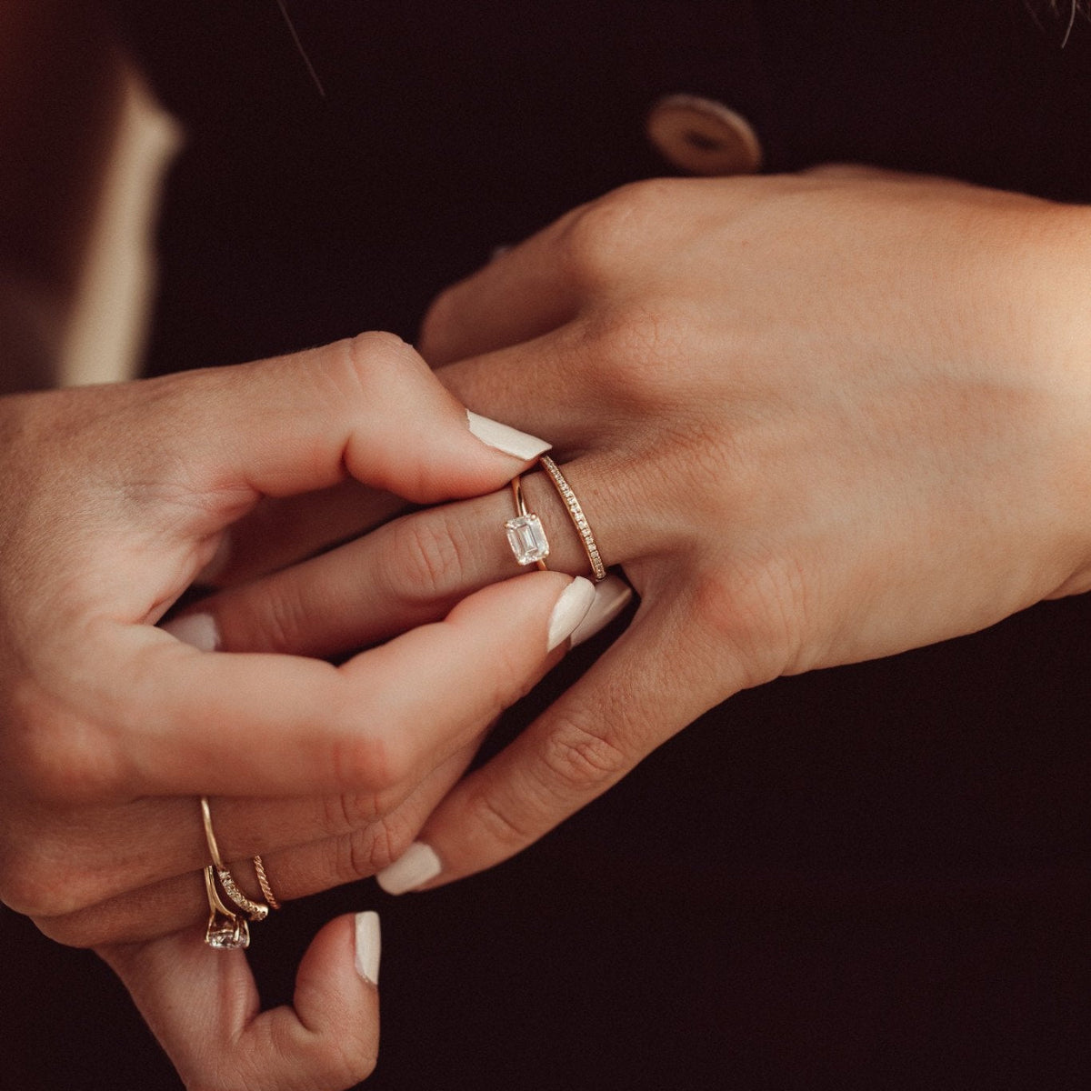 Close-up of hands wearing the Bennett emerald cut solitaire diamond ring and the single row diamond wedding band on a dark background.