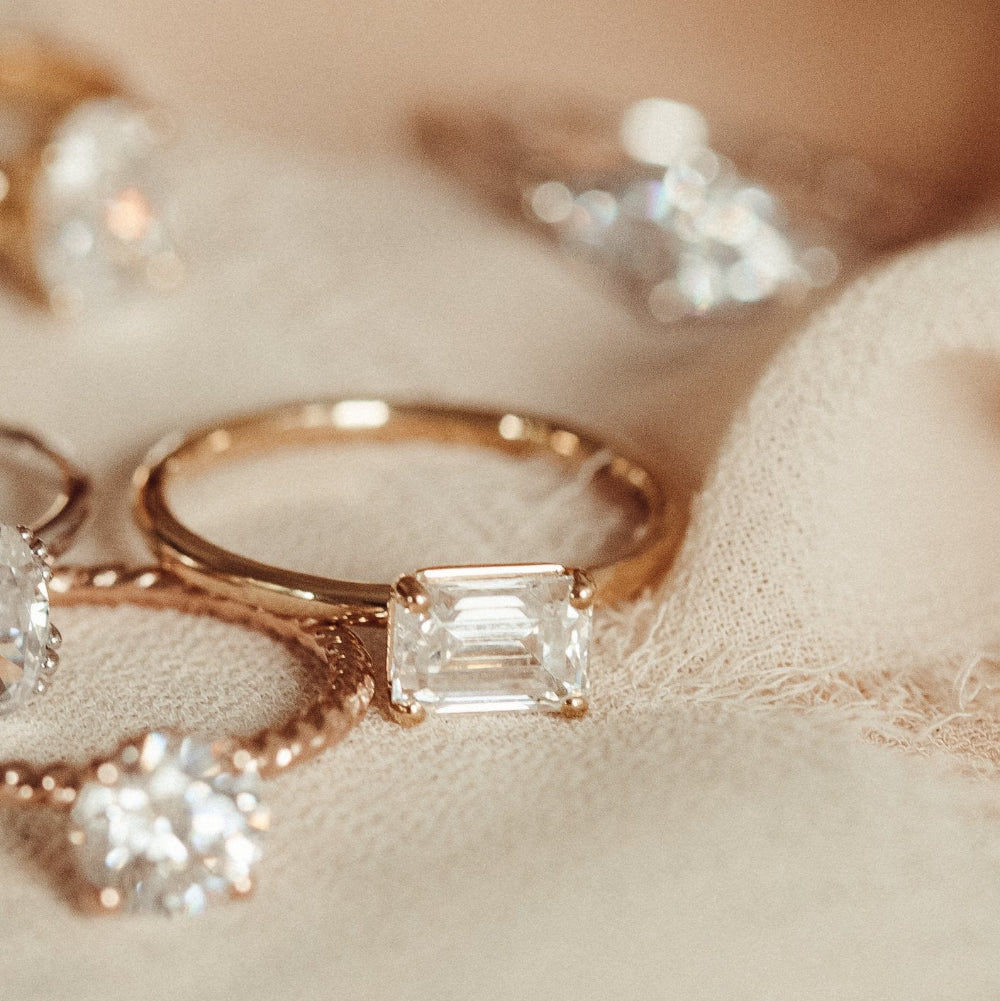 The Bennett emerald cut solitaire diamond ring on a beige fabric background