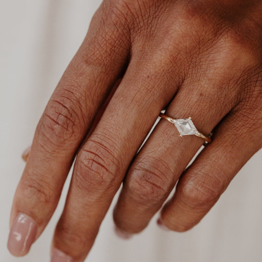 Hand wearing a lozenge diamond ring on a neutral background.