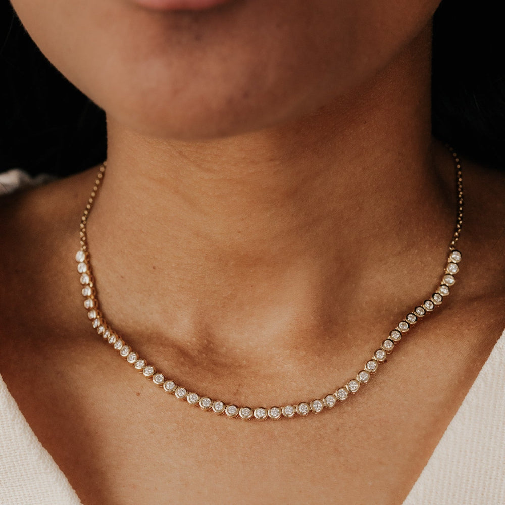 Close-up of a person wearing a delicate diamond tennis necklace on a dark background.