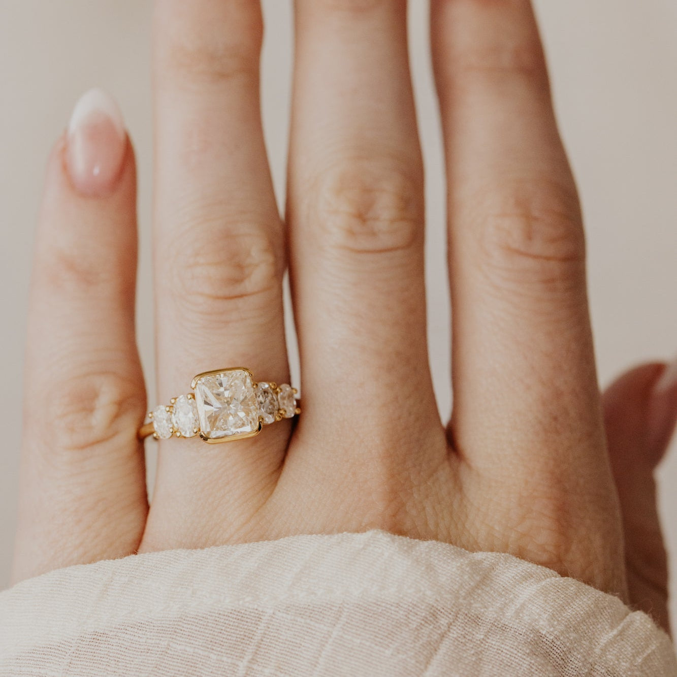 Hand wearing a gold ring with a cushion diamond on a neutral background.