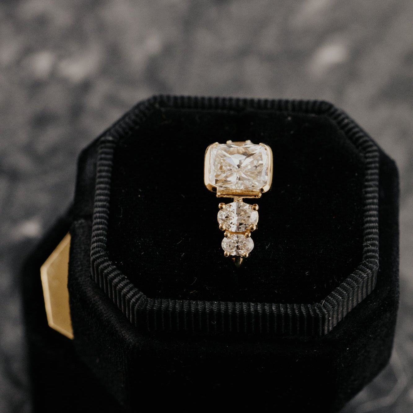 Gold ring with cushion diamond center stone and oval diamond side stones in a black velvet box on a textured surface.