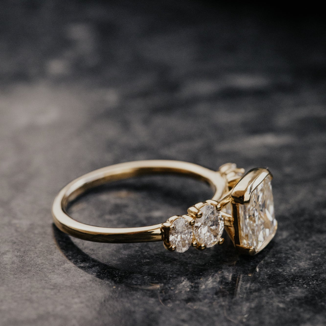 Gold ring with a cushion diamond center and oval diamond side stones on a dark surface.