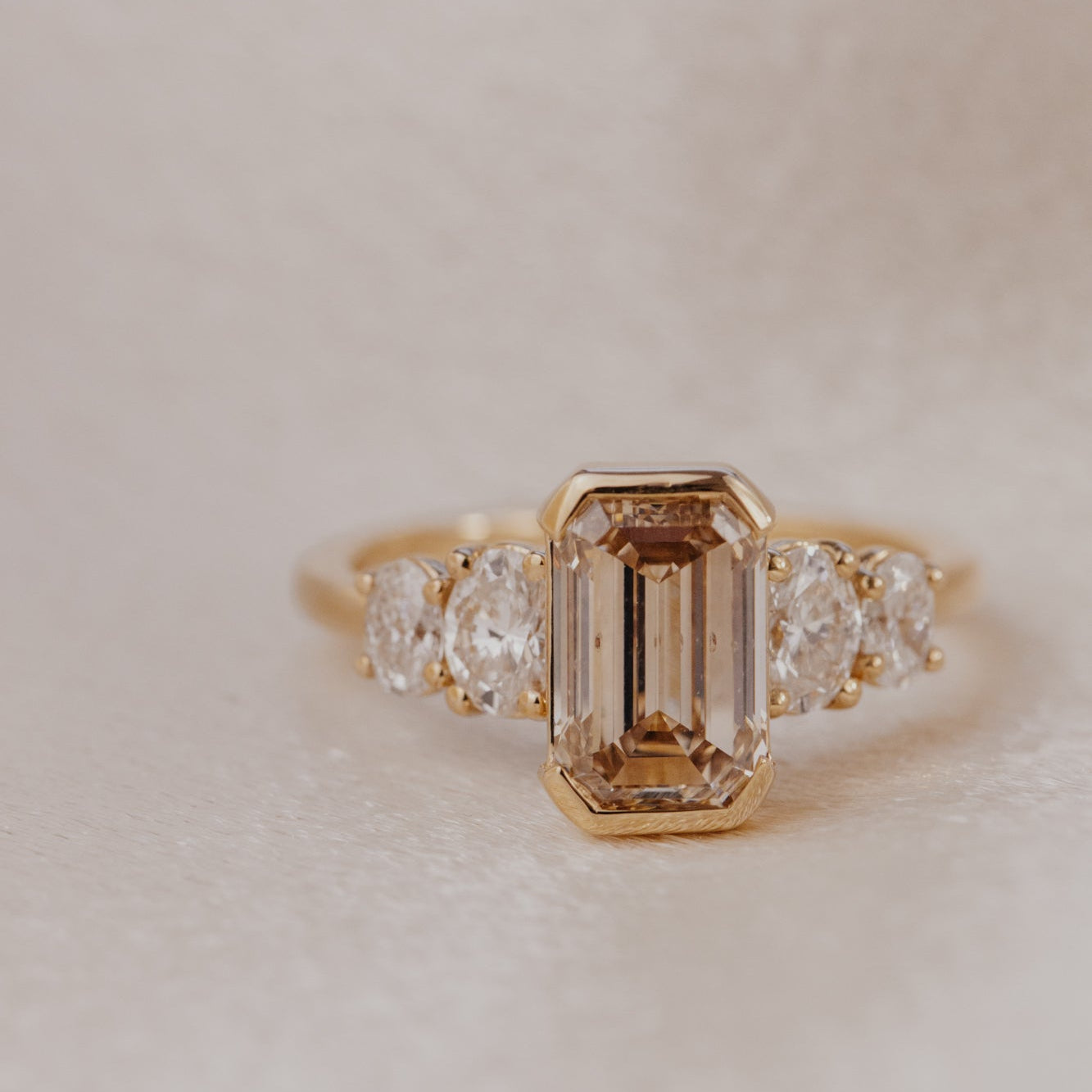 Gold ring with a central emerald cut champagne diamond and smaller stones on a light background.