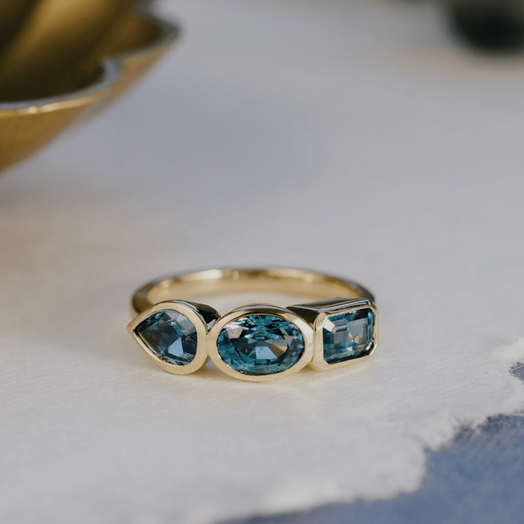 A 14k gold ring featuring three blue sapphire gemstones, displayed on a textured white surface with a soft-focus background.