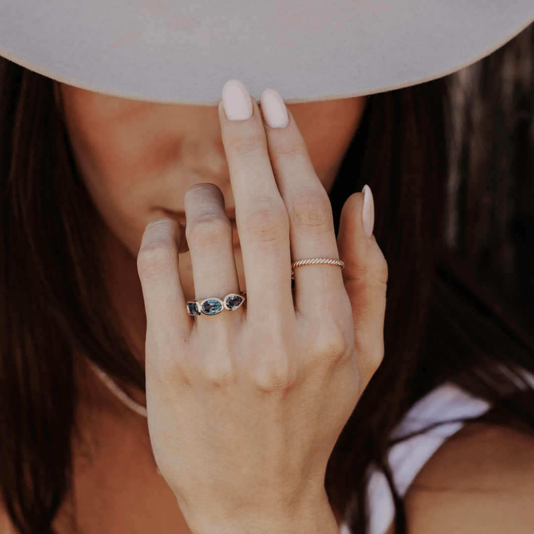 A close-up lifestyle shot of a woman's hand wearing two rings: a yellow gold skinny rope band and a yellow gold ring with three blue sapphire gemstones, while wearing a wide-brimmed light gray hat.