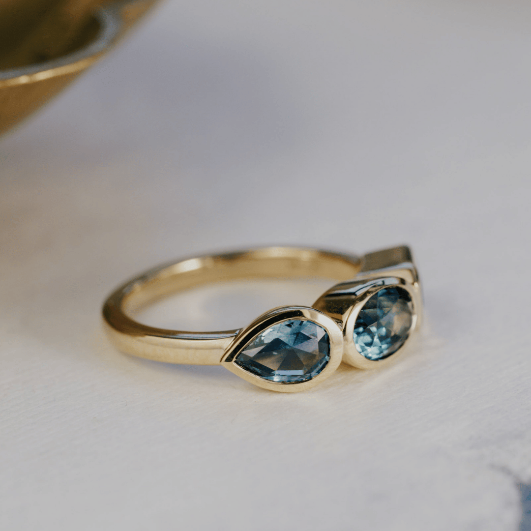 A yellow gold ring featuring a pear blue sapphire gemstones in a bezel setting, resting on a light textured surface.