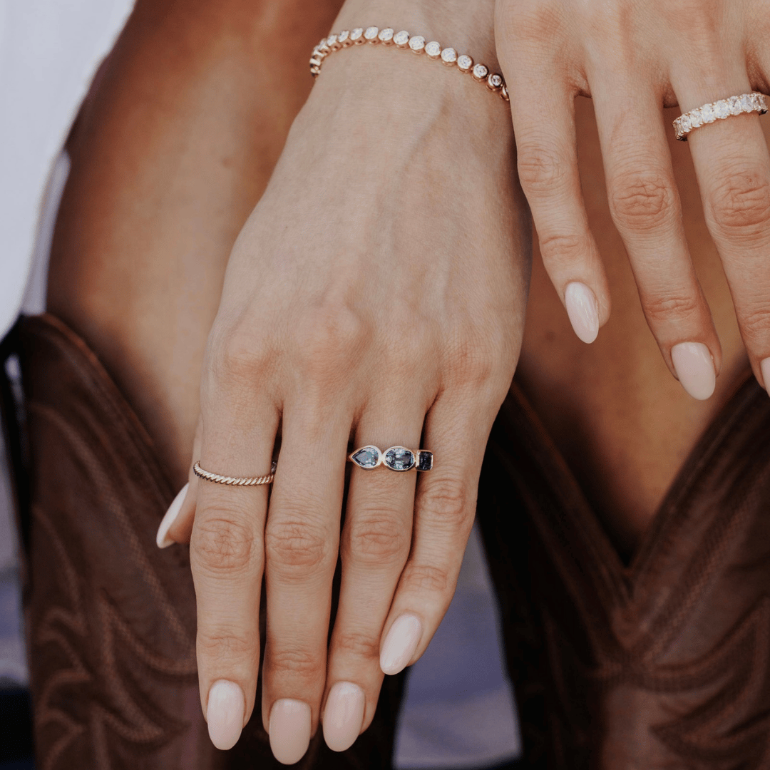 Close-up of a person's hands wearing multiple delicate gold rings with diamond and sapphire gemstones paired with a diamond tennis bracelet, with light pink manicured nails and brown western-style boots in the background.