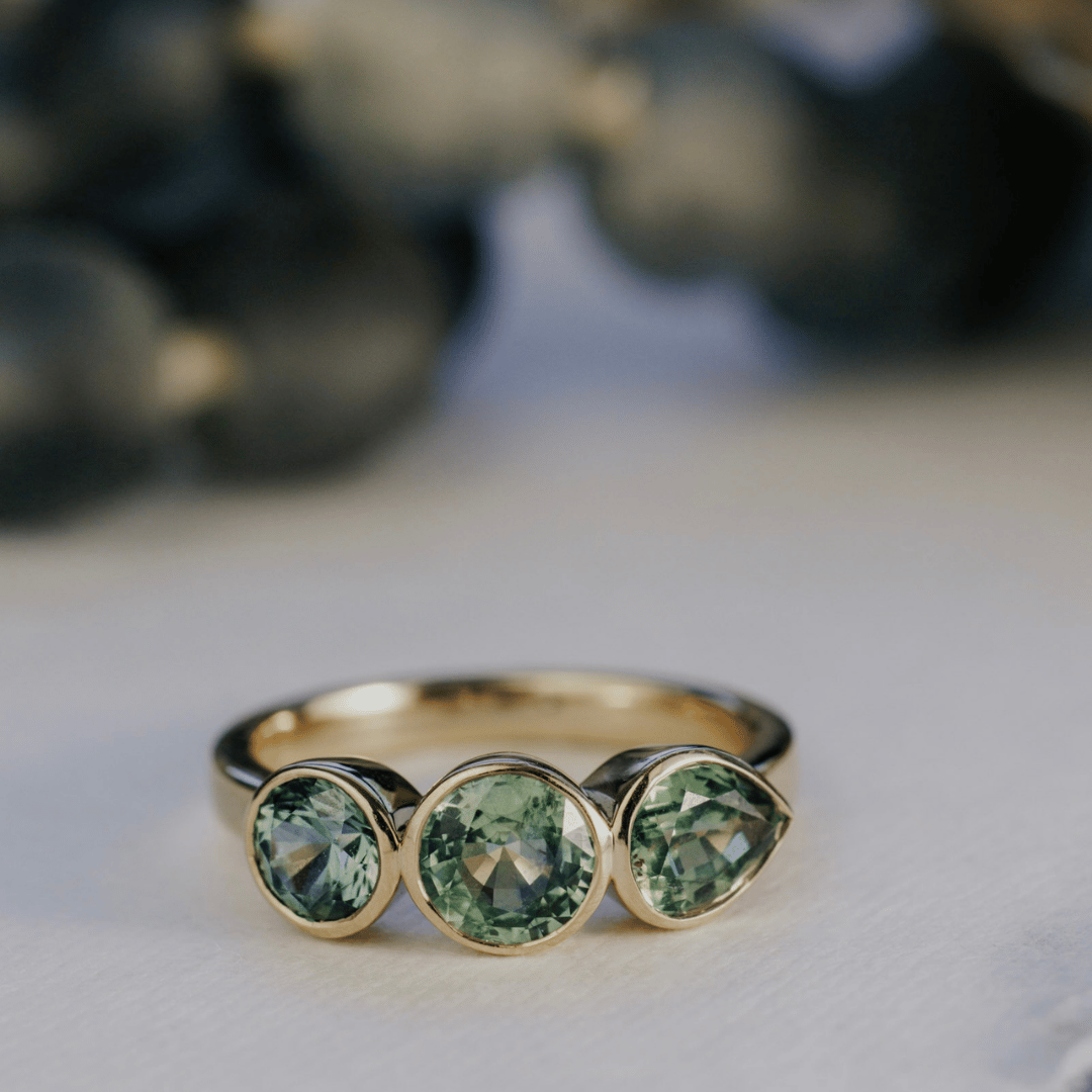 A 14k gold, ring featuring three blue sapphire gemstones, displayed on a textured white surface with a soft-focus background.
