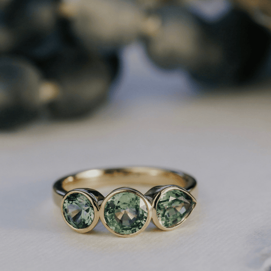 A 14k gold, ring featuring three blue sapphire gemstones, displayed on a textured white surface with a soft-focus background.
