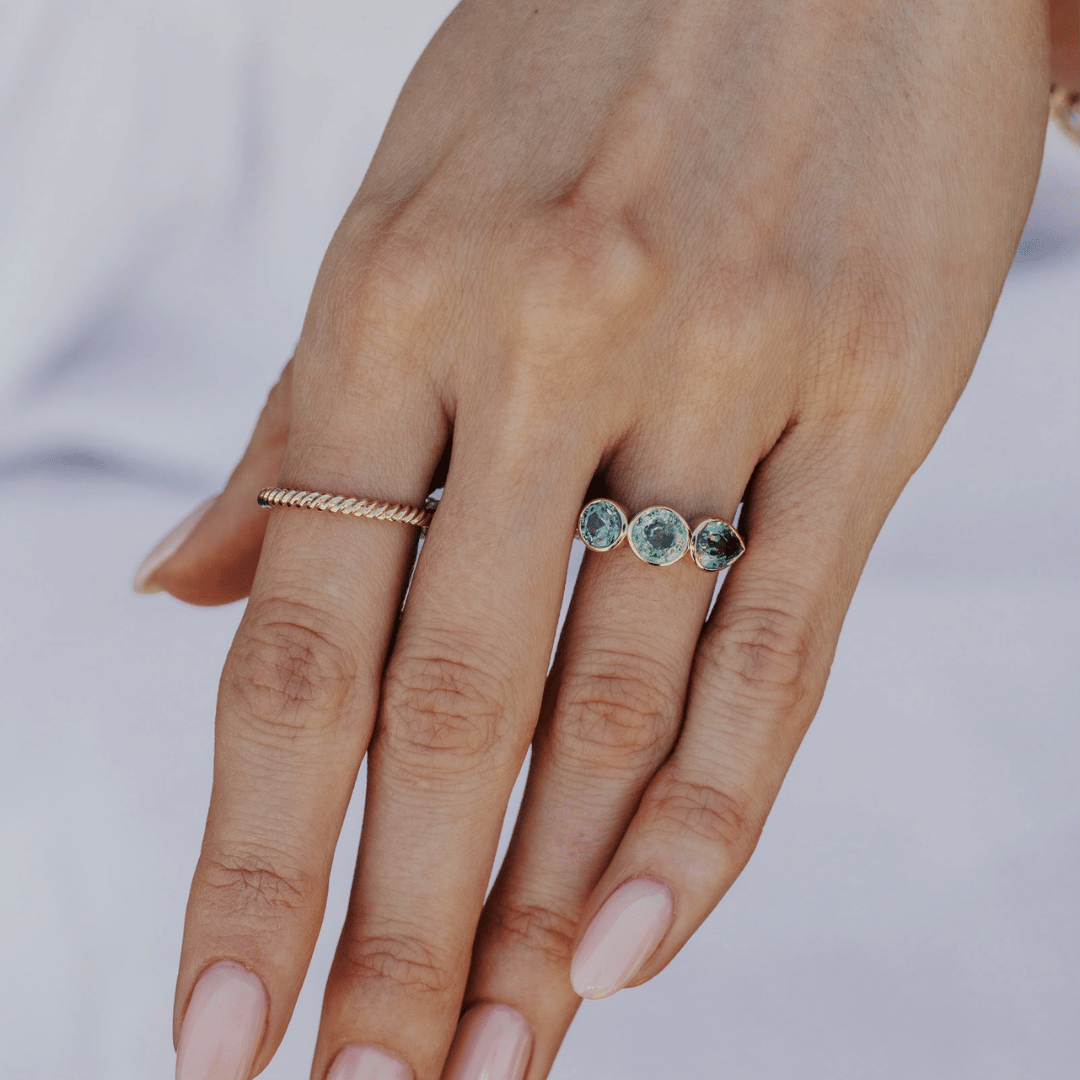 A 14k gold ring featuring three blue sapphire gemstones, displayed on the ring finger of a hand model with a soft-focus background.
