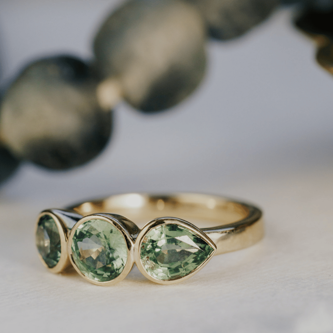 A 14k gold ring featuring three blue sapphire gemstones, displayed on a white surface angled to the side with the pear-shaped jewel as the image focus.
