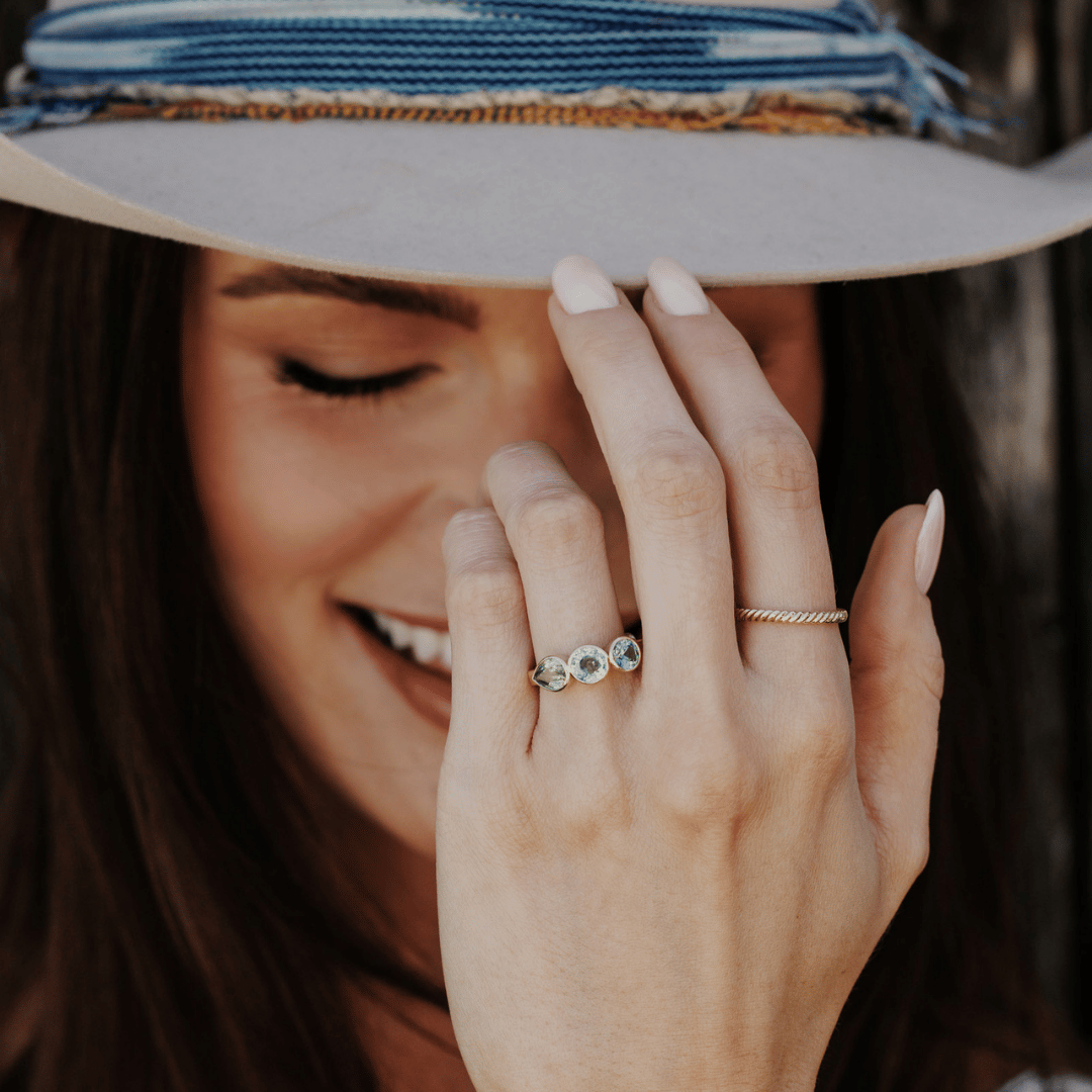 A 14k gold ring featuring three blue sapphire gemstones, displayed on the ring finger of a hand model touching the brim of her hat. 