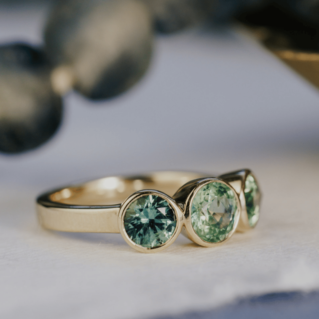 A 14k gold ring featuring three blue sapphire gemstones, displayed on a white surface angled to the side with the round-shaped jewel as the image focus.
